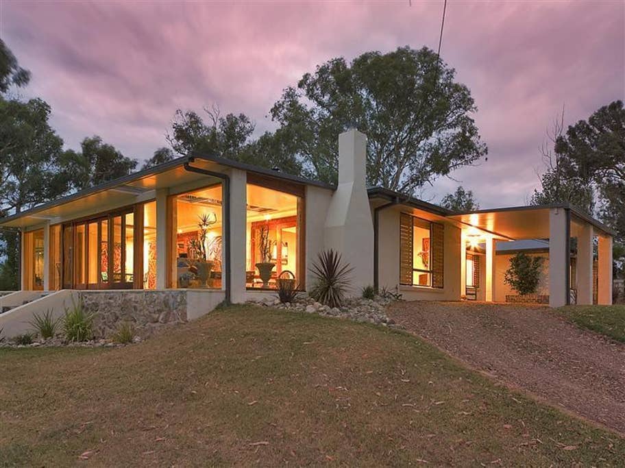 The modern single-story house is set on a grassy hill, featuring large glass doors that provide a view of the interior. The home is framed by lush trees and plantings, with a welcoming pathway leading to the entrance.
