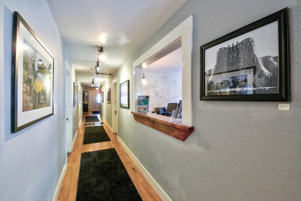 A long hallway is lined with framed artwork on light grey walls, featuring wooden trim. The wooden floor is complemented by a dark runner carpet. Light fixtures enhance the ambiance, and the hallway leads toward an inviting lobby area visible at the far end.