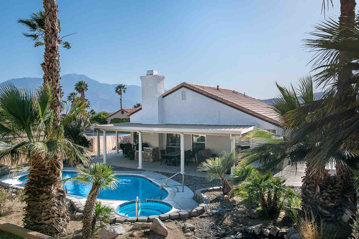 A spacious outdoor area features a swimming pool surrounded by tropical palm trees, with a separate spa nearby. A covered patio provides a shaded dining space, while the serene mountains serve as a scenic backdrop, enhancing the relaxed atmosphere of the property.