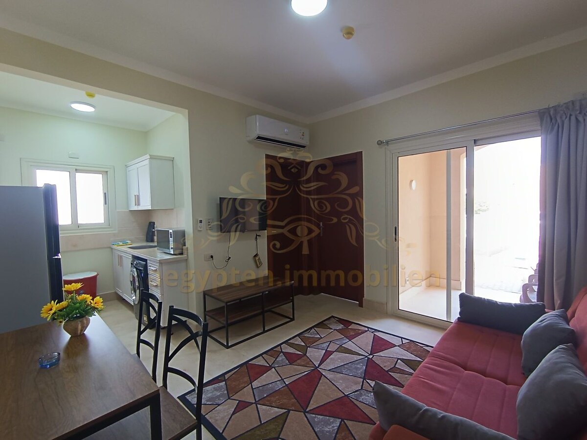 An open living area features a red sofa and a patterned area rug, complemented by a dining set and kitchen appliances. A door leads to an outdoor space, allowing natural light to fill the room. A small bouquet of flowers adds a touch of vibrancy.