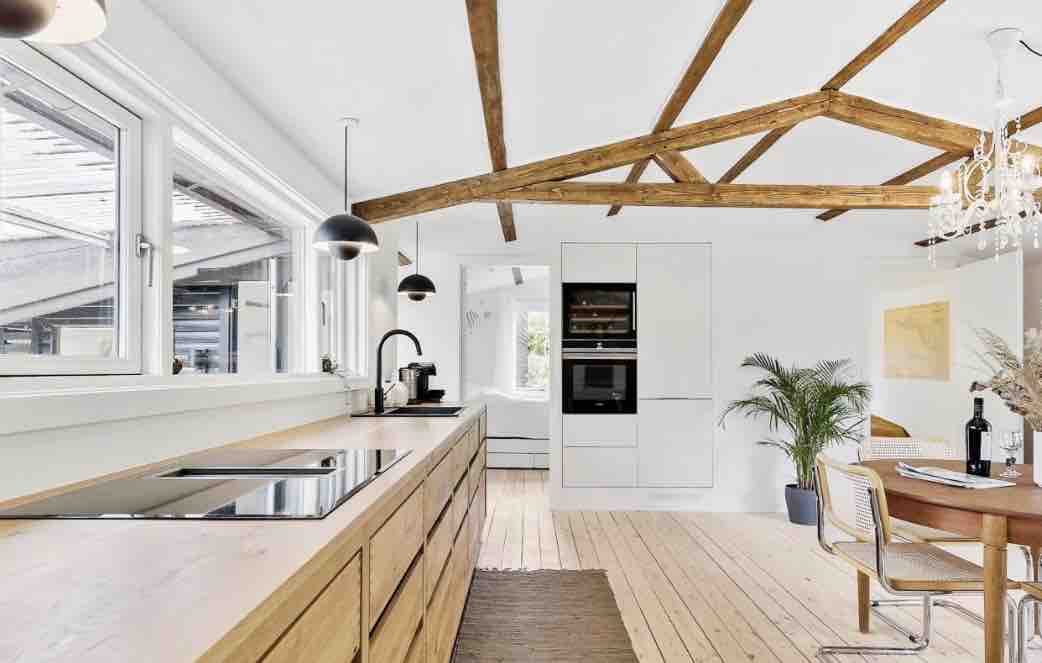 A spacious kitchen area features natural wood cabinets and a sleek countertop. Exposed wooden beams run across the ceiling, and two pendant lights hang above the workspace. Large windows allow natural light to fill the room, complemented by greenery in the corner and a dining table with stylish chairs.