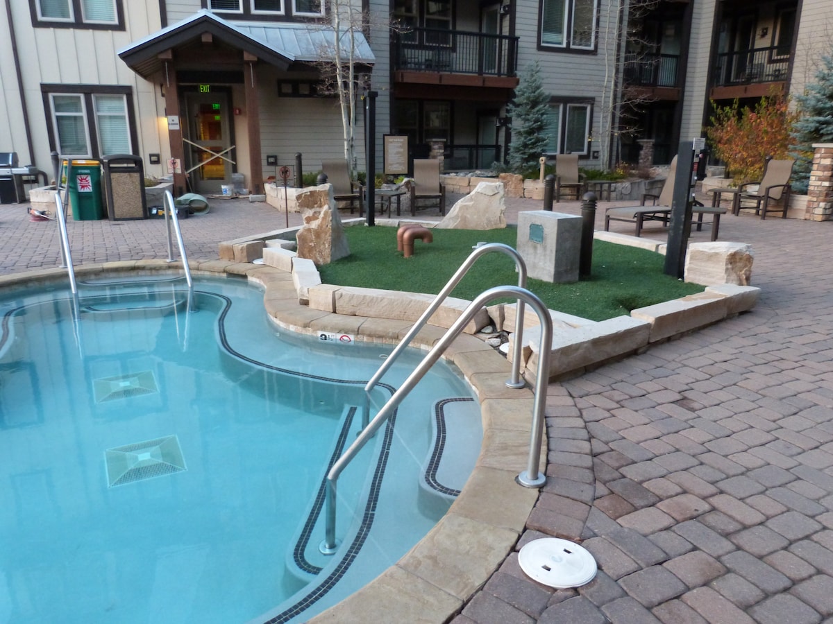 The outdoor amenity area features a clear swimming pool surrounded by a textured stone deck. Stainless steel handrails are positioned at the pool's entry, with landscaped sections of grass and rock formations nearby.