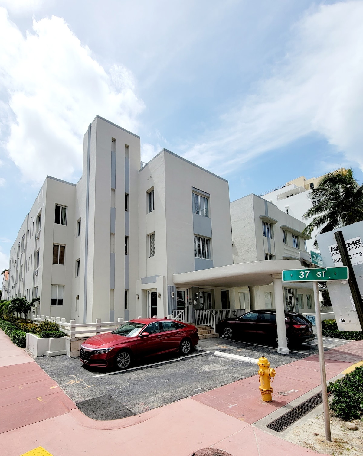 An art deco building is presented with a clean white facade, featuring large windows and geometric lines. Two cars are parked in front, while a street sign indicates the nearby intersection. Lush greenery borders the driveway and sidewalk, contributing to the welcoming exterior.