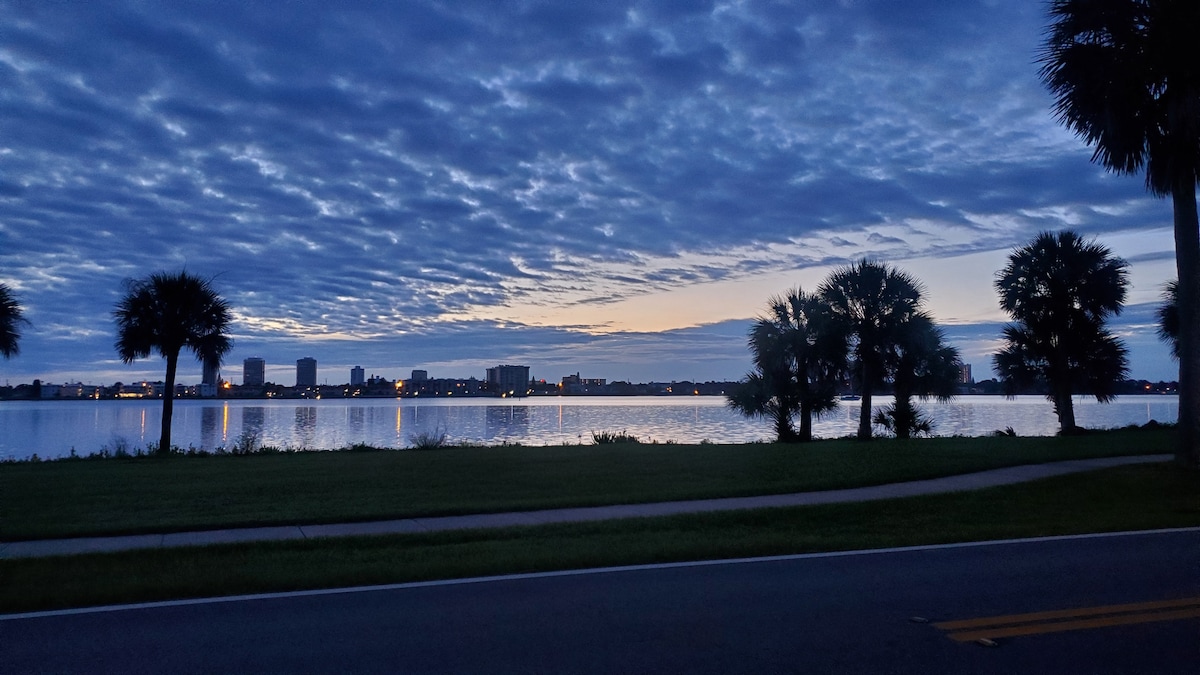 A serene body of water reflects the soft blue hues of dawn, with silhouettes of palm trees framing the foreground. The skyline features buildings highlighted by early morning lights, creating a peaceful atmosphere as clouds drift above.