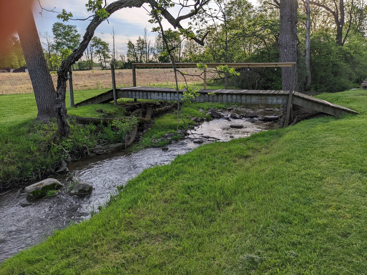 A peaceful stream flows gently through the property, bordered by lush green grass. A wooden bridge crosses over the water, supported by sturdy beams. Surrounding trees provide shade and add to the serene atmosphere of the natural setting.
