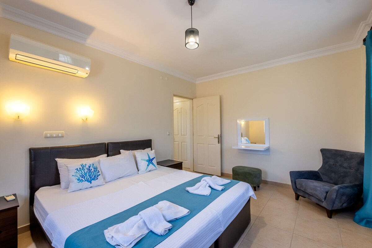 A spacious sleeping area is furnished with a large bed, complemented by crisp white linens and teal accents. Soft lighting is provided by wall sconces and a ceiling fixture. A wall-mounted mirror and a cozy armchair are also present, contributing to the room's functional design.