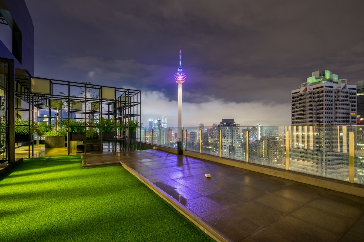 The rooftop garden features a well-maintained green area with artificial grass, surrounded by modern structures and planters. A clear view of the city skyline is provided, including a prominent tower illuminated with colorful lights against the night sky.