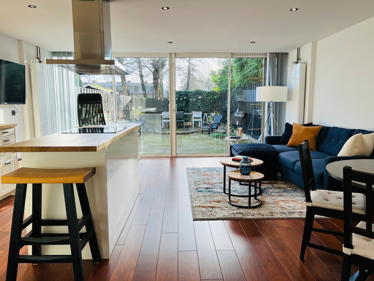 A spacious living area is highlighted by large glass doors, allowing natural light to brighten the room. A cozy couch is arranged with decorative pillows, and a stylish rug complements the wooden flooring. An open kitchen features a central island and modern appliances.