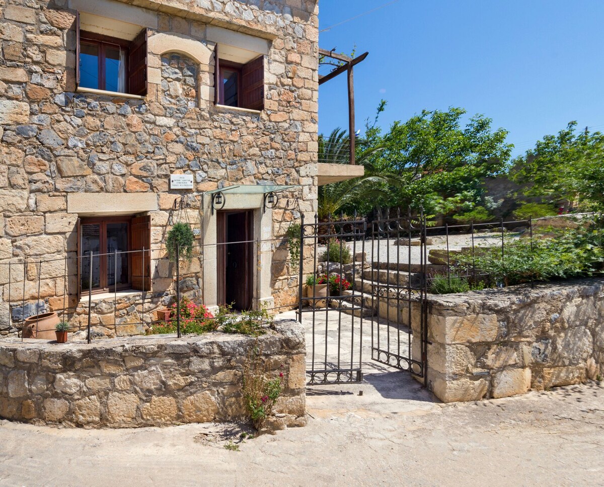The exterior of the stone cottage is displayed, showcasing traditional architecture with wooden window shutters. A decorative gate and small garden area with potted plants are visible, while the surrounding landscape features greenery and a clear blue sky.