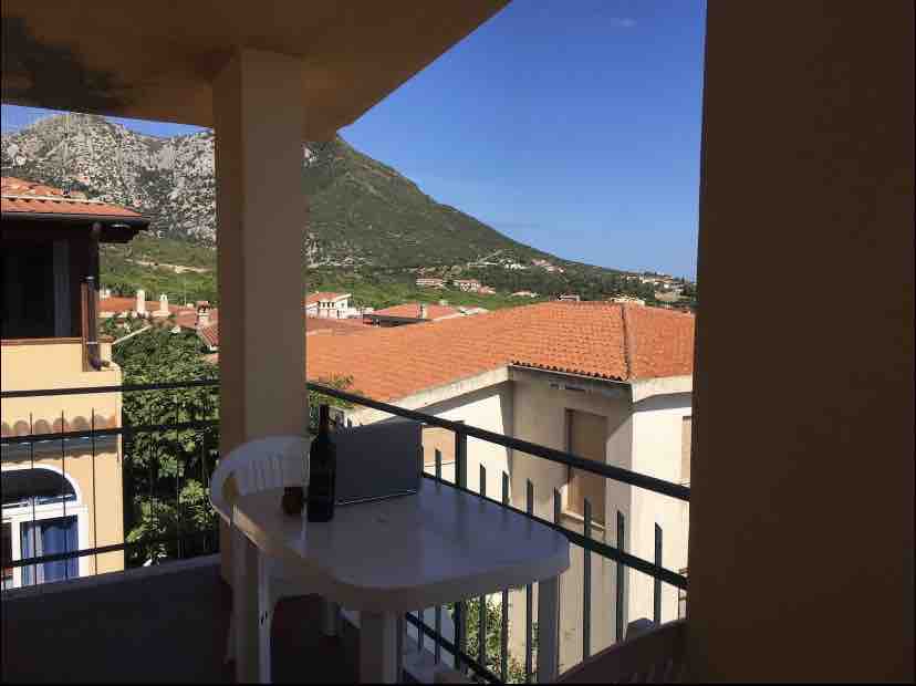 A balcony features a small table and two white chairs, positioned to enjoy the scenic view of rolling hills and rooftops. The blue sky presents a clear backdrop, while the surrounding landscape showcases greenery and terracotta-tiled roofs.