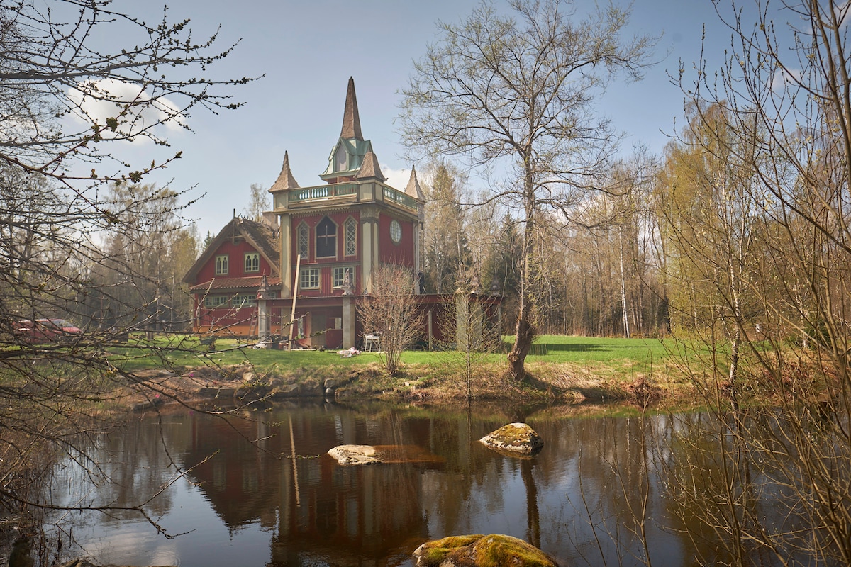 A whimsical house with a distinct spire stands beside a serene body of water, surrounded by trees and open green spaces. The structure features a multi-level design, offering an inviting presence in a tranquil natural setting.