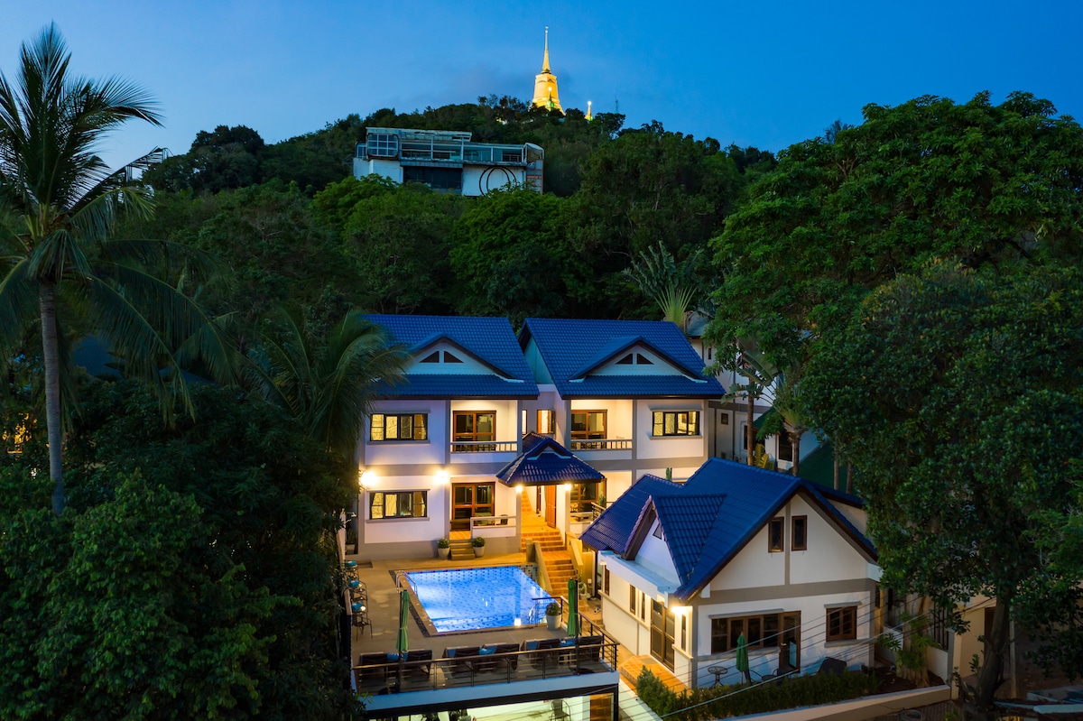 A serene view of a multi-story property nestled among lush greenery, featuring a sparkling pool at the center. A illuminated Buddha statue is visible on the hillside in the background, providing a peaceful ambiance during twilight hours.