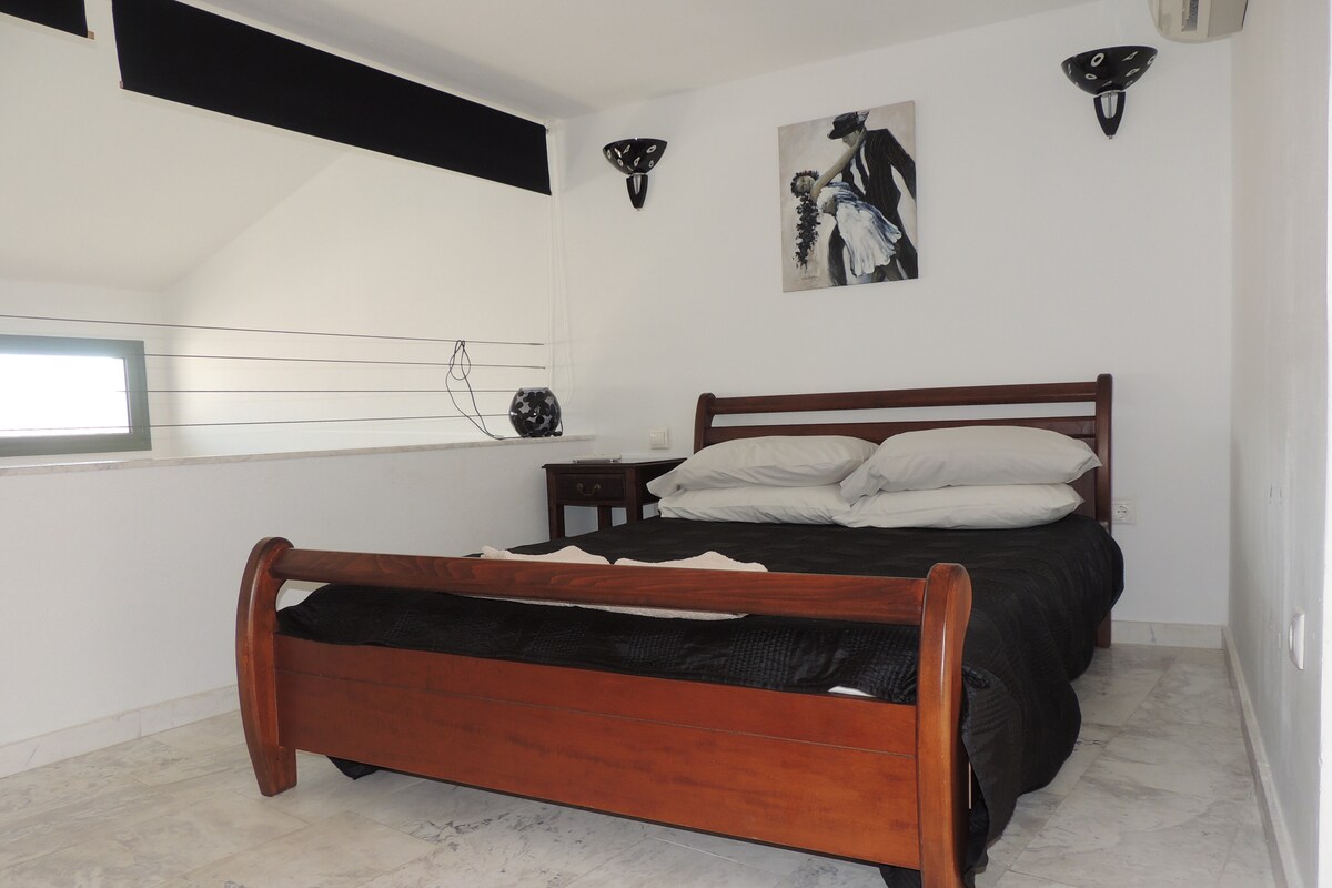 A modern bedroom features a wooden bed with neutral-toned bedding. Two wall sconces provide soft lighting above the bed, complemented by a piece of wall art. A nightstand is positioned beside the bed, and natural light is diffused through a nearby window.