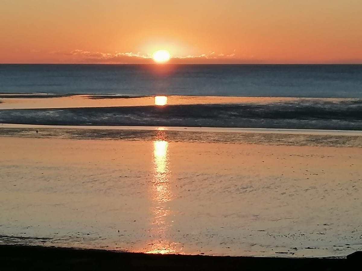 A serene sunset is captured over the calm sea, with the sun reflecting on the water's surface. Hues of orange and pink blend in the sky, while the shoreline is softly illuminated. The scene evokes tranquility and natural beauty.
