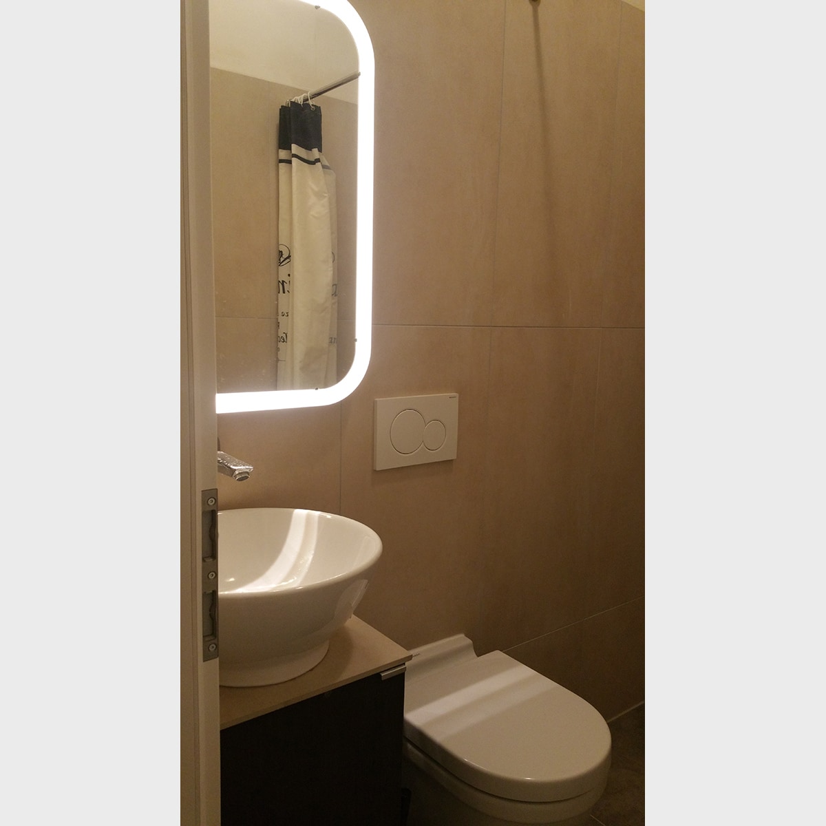 A modern bathroom is featured, showcasing a wall-mounted mirror with integrated lighting. A round sink is set on a minimalistic counter, complemented by a stylish faucet. The toilet is positioned beside the sink, with neutral-toned tiles enveloping the space.