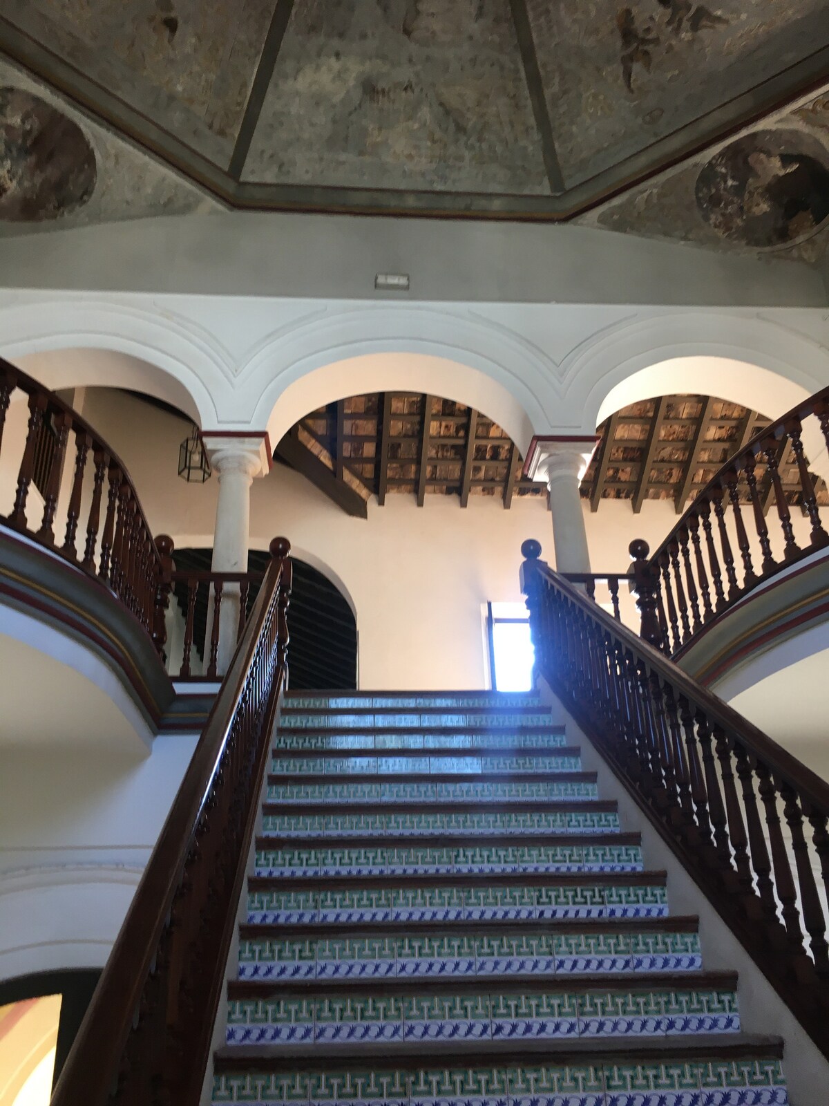 An elegantly designed staircase is visible, leading to an upper level. Intricate wooden railings frame the stairs, while decorative tile work lines the steps. The ceiling features ornate detailing, and natural light enters through windows at the top, illuminating the architectural features.