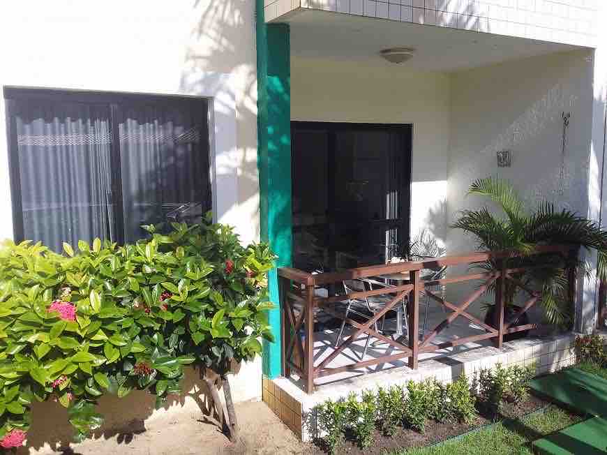 A shaded balcony is viewed, featuring a wooden railing and a table with several chairs. Lush greenery surrounds the space, creating a pleasant environment. The entrance to the apartment is visible, leading into the interior area with natural light filtering through the sliding glass doors.