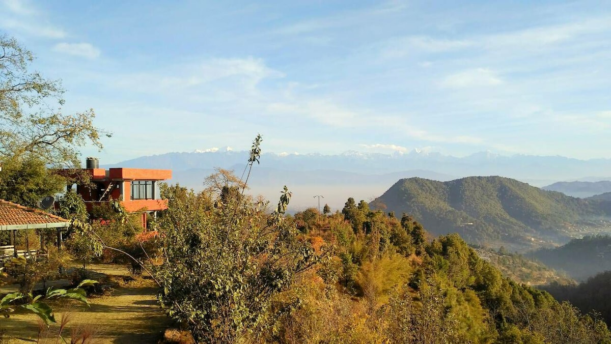 The image captures a panoramic view of the Himalayas, framed by lush greenery and sloping hills. A red building, part of the resort, is visible alongside the natural landscape, which includes various trees and vegetation, all under a clear, blue sky.