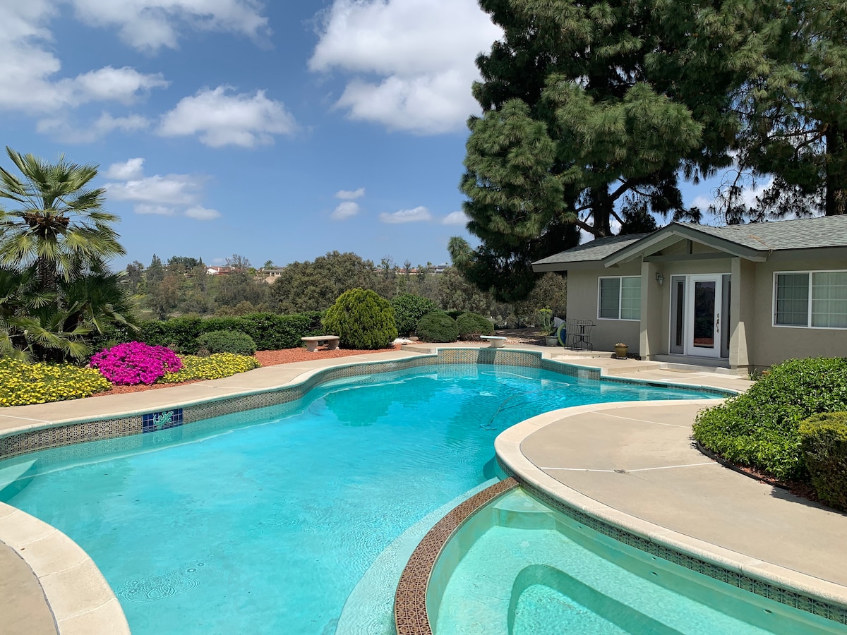 A sparkling pool is surrounded by lush greenery and colorful flower beds, providing a serene outdoor space. The guesthouse is visible in the background, with access to the patio area and the inviting blue water reflecting the clear sky.