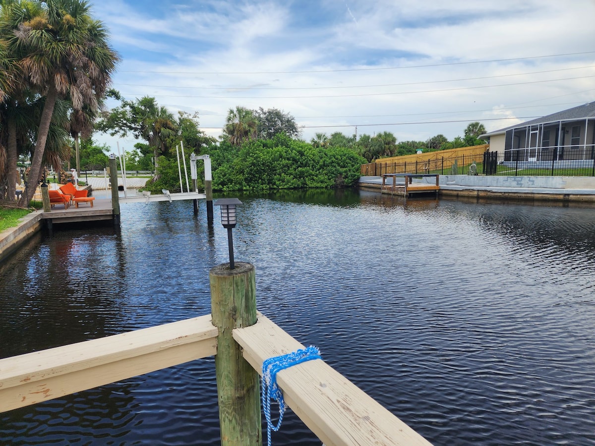 A serene view of the calm waterway features a wooden dock with a boat lift and a cozy outdoor seating area. Lush palm trees border the scene, adding greenery, while a neighboring property is visible across the water. The sky is partly cloudy, enhancing the tranquil ambiance.