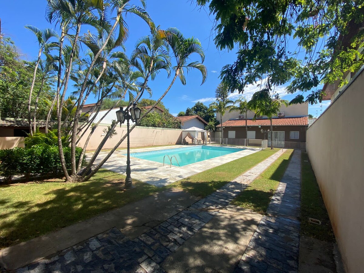 A serene outdoor area features a large swimming pool surrounded by a paved terrace and lush greenery. Palm trees provide shade along the pathway, leading to the pool area. A small garden with grass complements the space, ideal for relaxing.