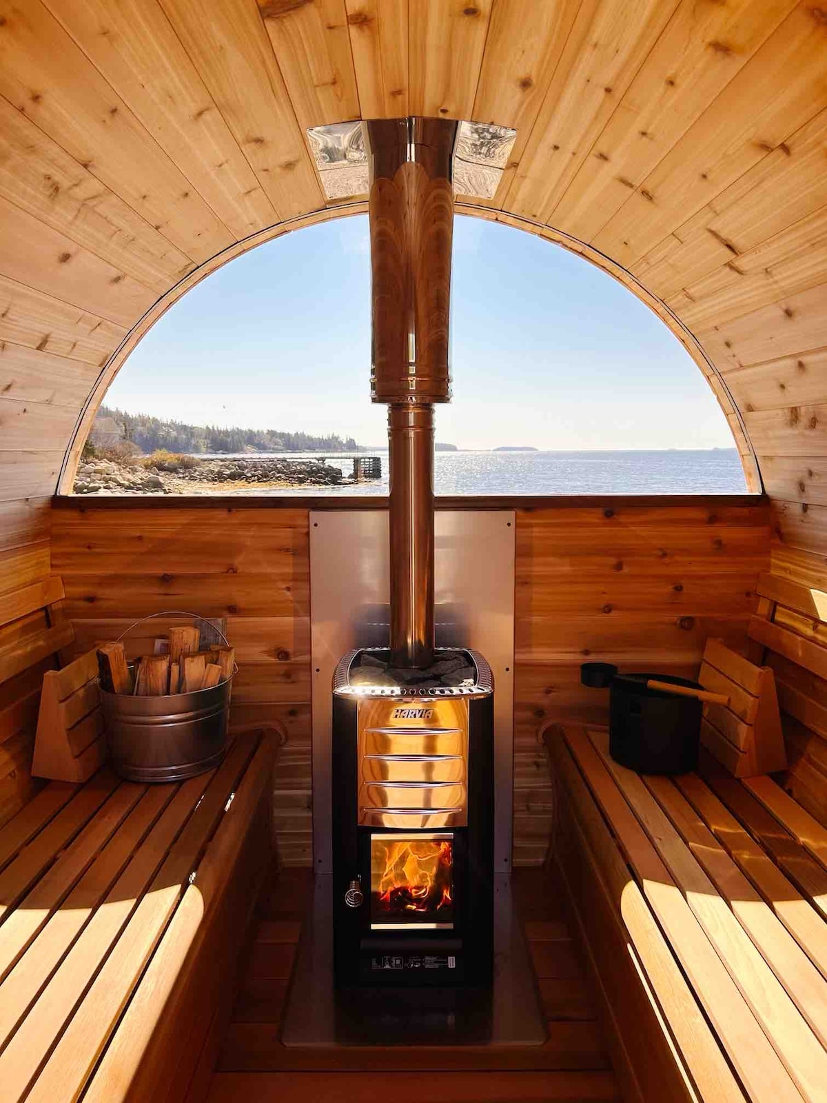 The interior of the wood-fired sauna features wooden bench seating along the curved walls and a stove with a chimney at the center. Large windows provide a view of the ocean, allowing natural light to brighten the space.