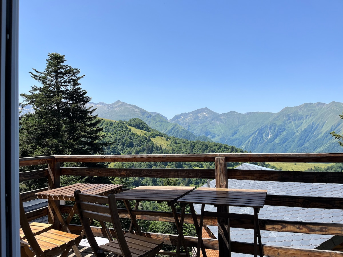 A wooden balcony offers sweeping views of rolling green mountains under a clear blue sky. The outdoor seating area includes a table and folding chairs, inviting relaxation amidst a serene natural landscape.