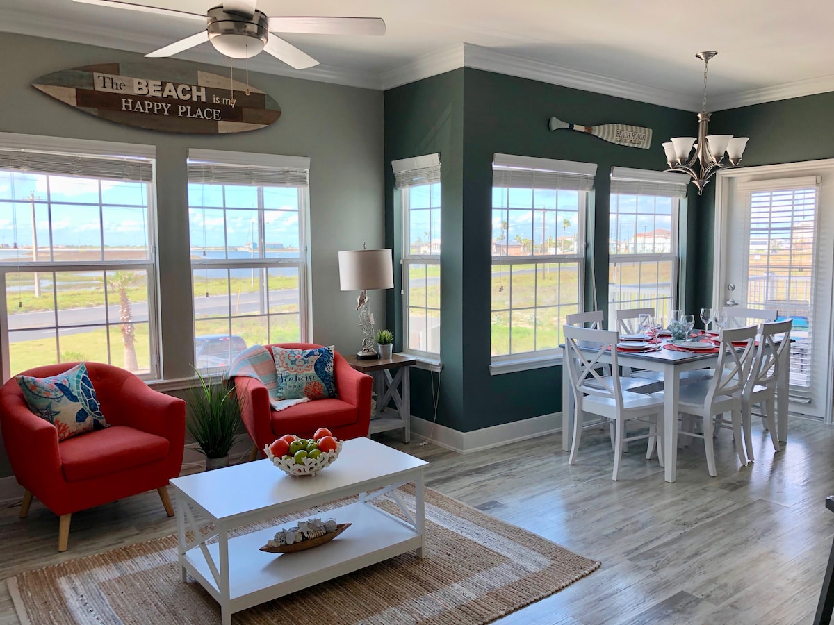 A bright and airy living space features large windows, allowing natural light to fill the room. Two red chairs are positioned beside a white coffee table, while a dining area with a white table and chairs is visible. Beach-themed decor accents the walls, enhancing the coastal vibe.