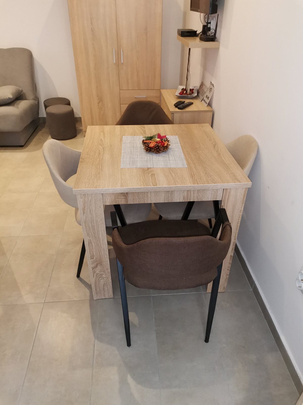 A small dining area features a wooden table surrounded by four chairs. A centerpiece with decorative flowers rests on the table. Light-colored tiles cover the floor, and a piece of furniture is visible in the background.