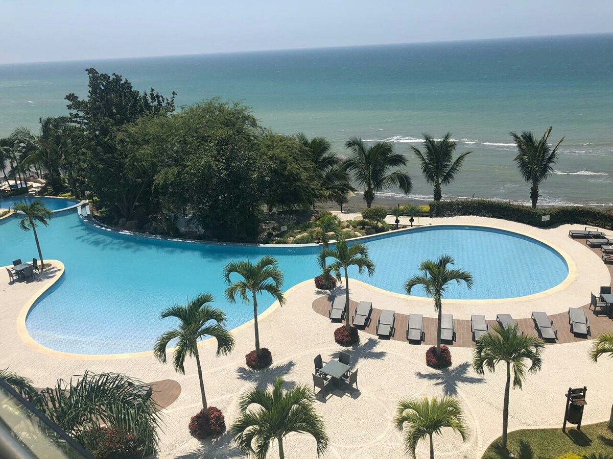 A spacious outdoor pool area is surrounded by lush palm trees and well-maintained gardens. The inviting blue waters of the pool reflect the sunlight, while the ocean is visible in the background, offering a serene coastal view.