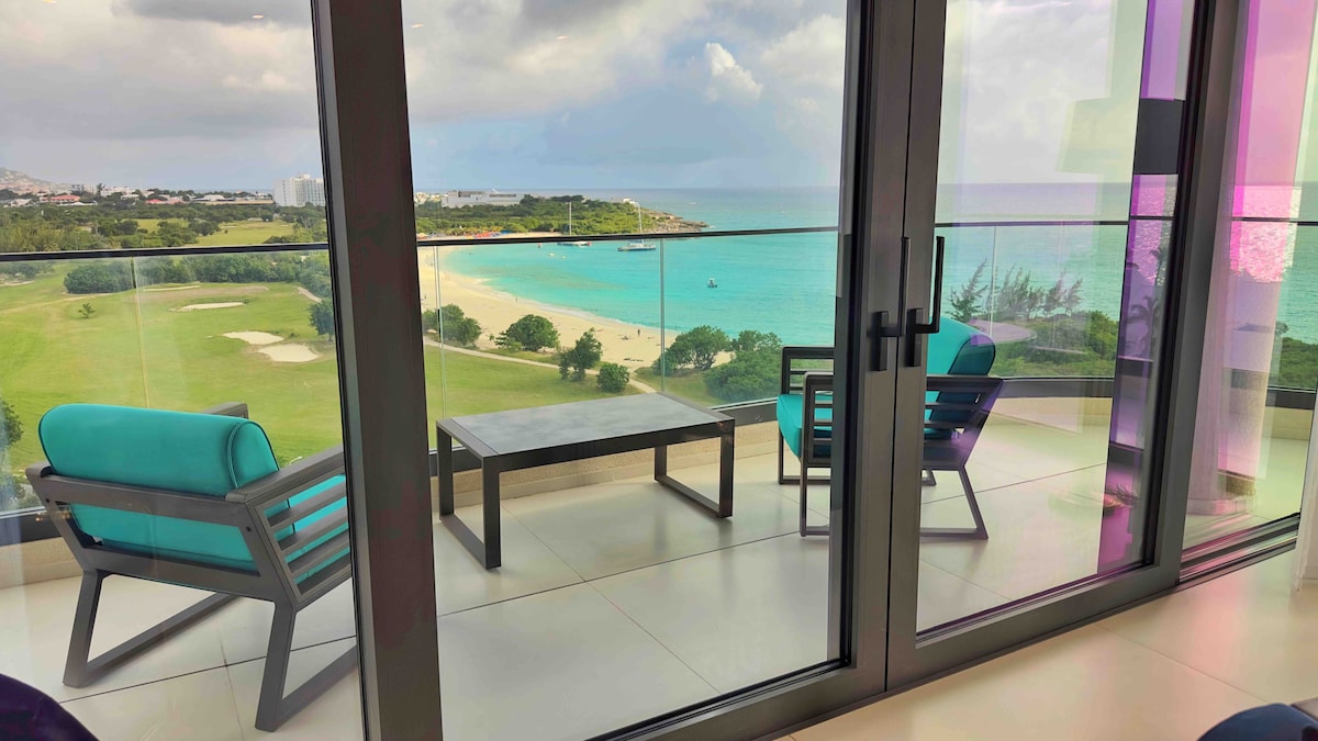 A balcony is displayed featuring two modern chairs with teal cushions and a small table, overlooking a panoramic view of a lush green golf course and the turquoise ocean. The scene captures a tranquil atmosphere with the beach in the distance.