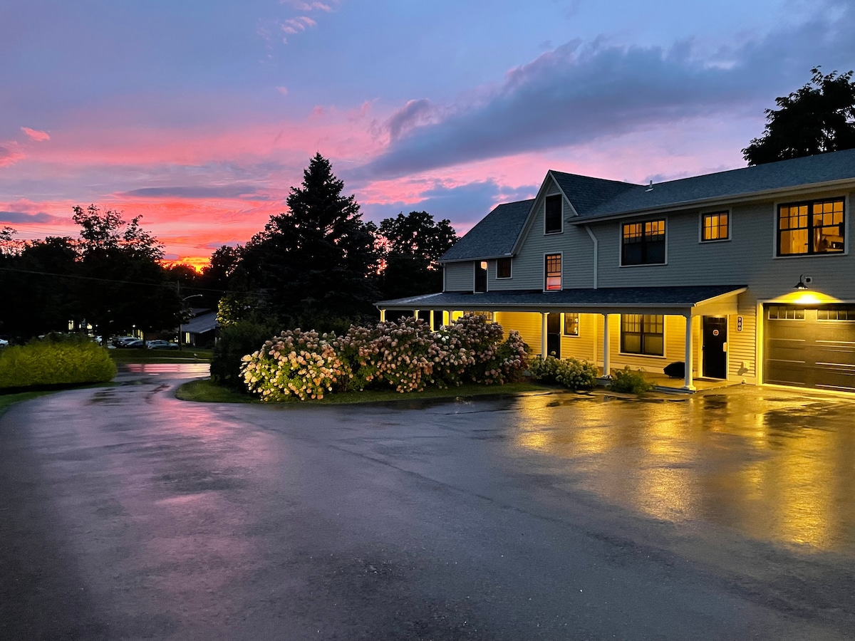 An evening view showcases the exterior of the home, with vibrant sunset colors reflected in the wet driveway. Lush bushes filled with blooming flowers line the parking area, while the house structure stands under a dramatic sky framed by trees.
