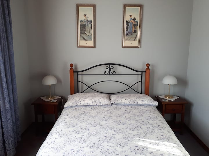 A classic bedroom with a bed featuring floral-patterned bedding and a metal headboard with wooden posts. Two bedside tables hold matching white lamps, and two framed art pieces hang above the bed. The room has a simple, traditional style with blue curtains.