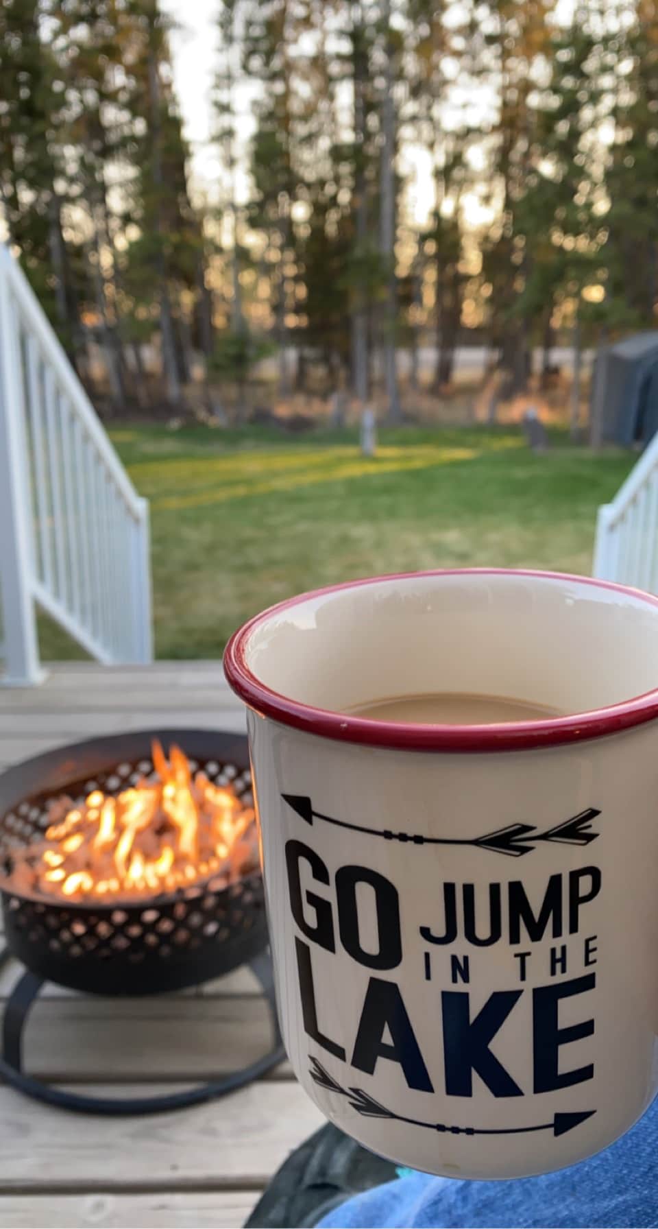 A warm beverage is held in a cup with a playful message. In the background, a fire pit glows with flickering flames, and lush trees frame the serene yard, creating a peaceful outdoor setting.