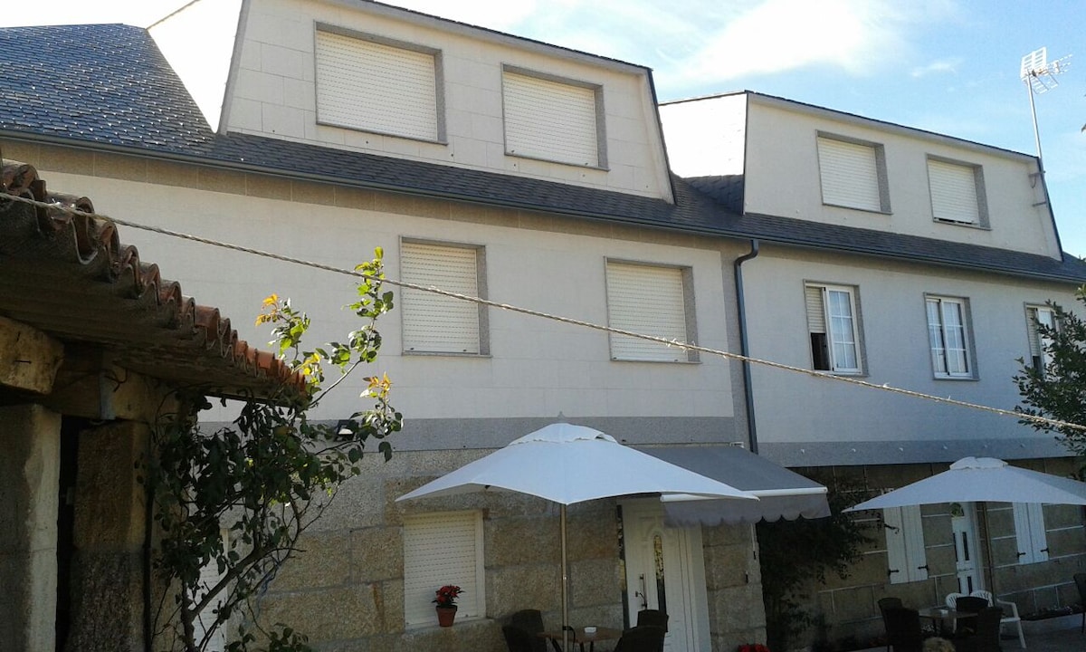 The exterior of the building features white stone walls and a sloped grey roof. Window shutters are closed, and two large umbrellas provide shade over an outdoor seating area with chairs. A small flowering plant adds a touch of greenery to the scene.