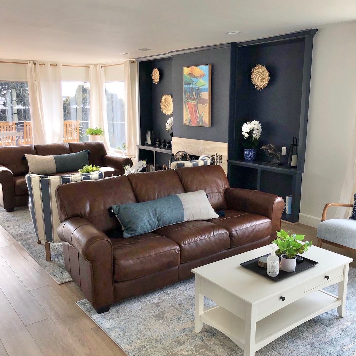 A spacious living area is highlighted by a brown leather sofa and striped armchairs. A modern coffee table with decorative items sits in the center. Natural light filters through large windows, illuminating artwork on the wall and potted plants arranged throughout the room.