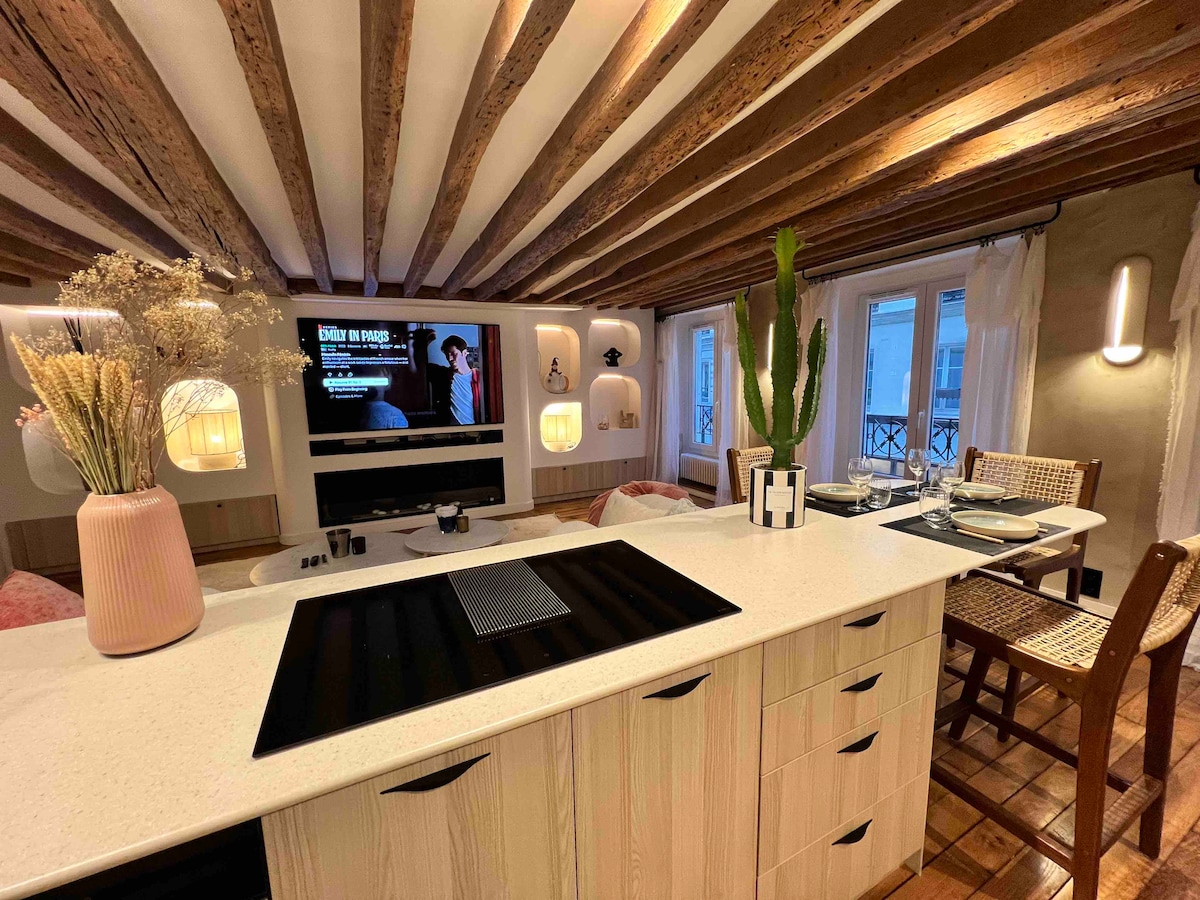 A modern kitchen showcases a sleek countertop with an induction cooktop and ample cabinetry. A flat-screen TV is mounted on the wall, enhancing entertainment options. Decorative plants and a vase of dried flowers add a touch of warmth. Wooden beams create visual interest overhead.