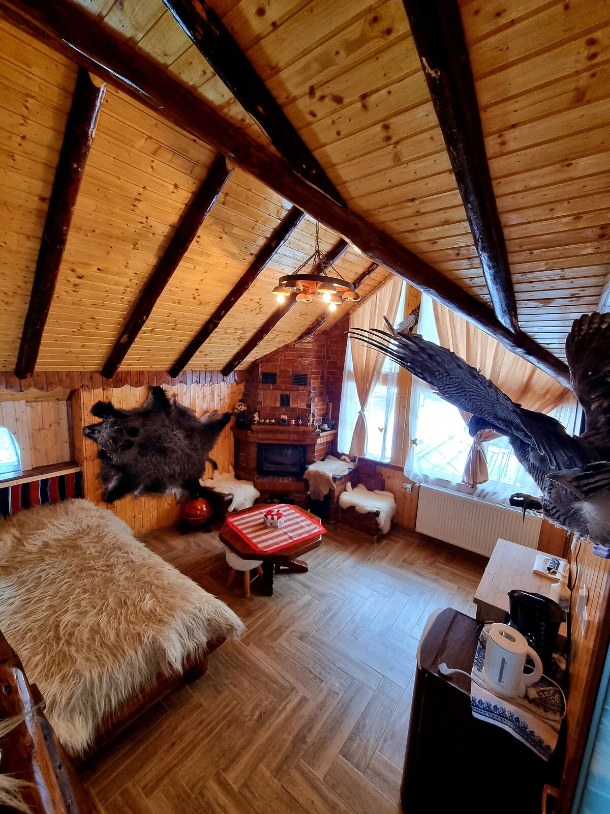 The interior features a wooden ceiling with exposed beams and rustic decor. A cozy seating area includes a daybed with a fluffy throw, a small table, and a traditional wood-burning stove. Natural light enters through the windows covered by light curtains, adding warmth to the space.