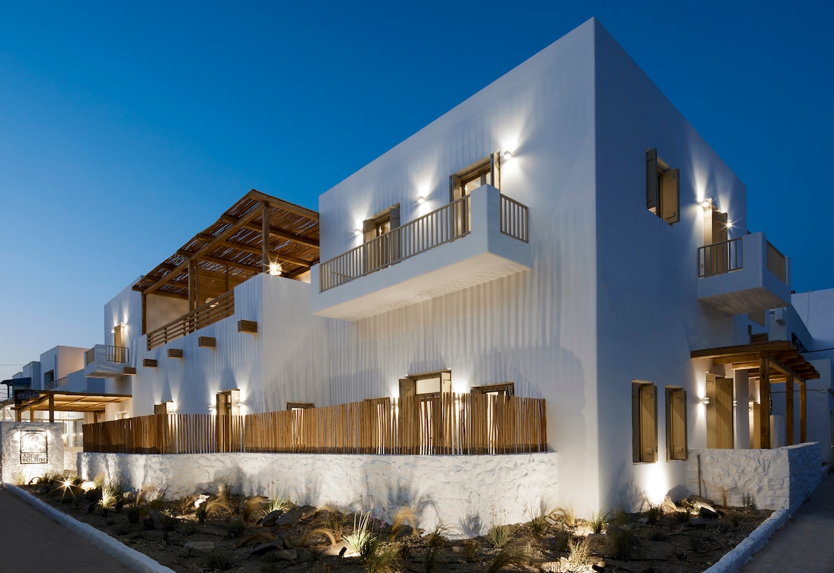 The exterior features a modern building, characterized by smooth white walls and wooden accents. Soft lighting highlights architectural details, while the multi-level structure includes spacious balconies. Natural landscaping elements complement the design, creating an inviting atmosphere under the evening sky.