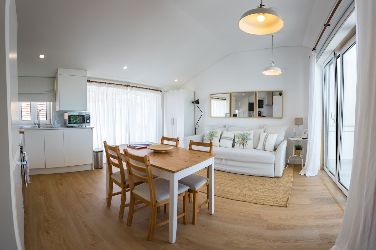A bright living area features large windows allowing natural light to fill the space. A dining table with four chairs is positioned near the kitchen. A comfortable white sofa is accessorized with patterned cushions, complemented by a simple area rug. Modern light fixtures hang from the ceiling.