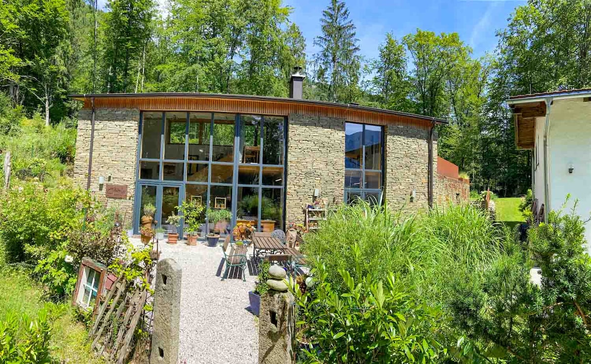 The exterior of the loft features a stone facade with large glass windows allowing abundant natural light. A spacious patio is surrounded by greenery and landscaped with potted plants, offering a serene retreat. The setting conveys a harmonious blend of nature and architecture.