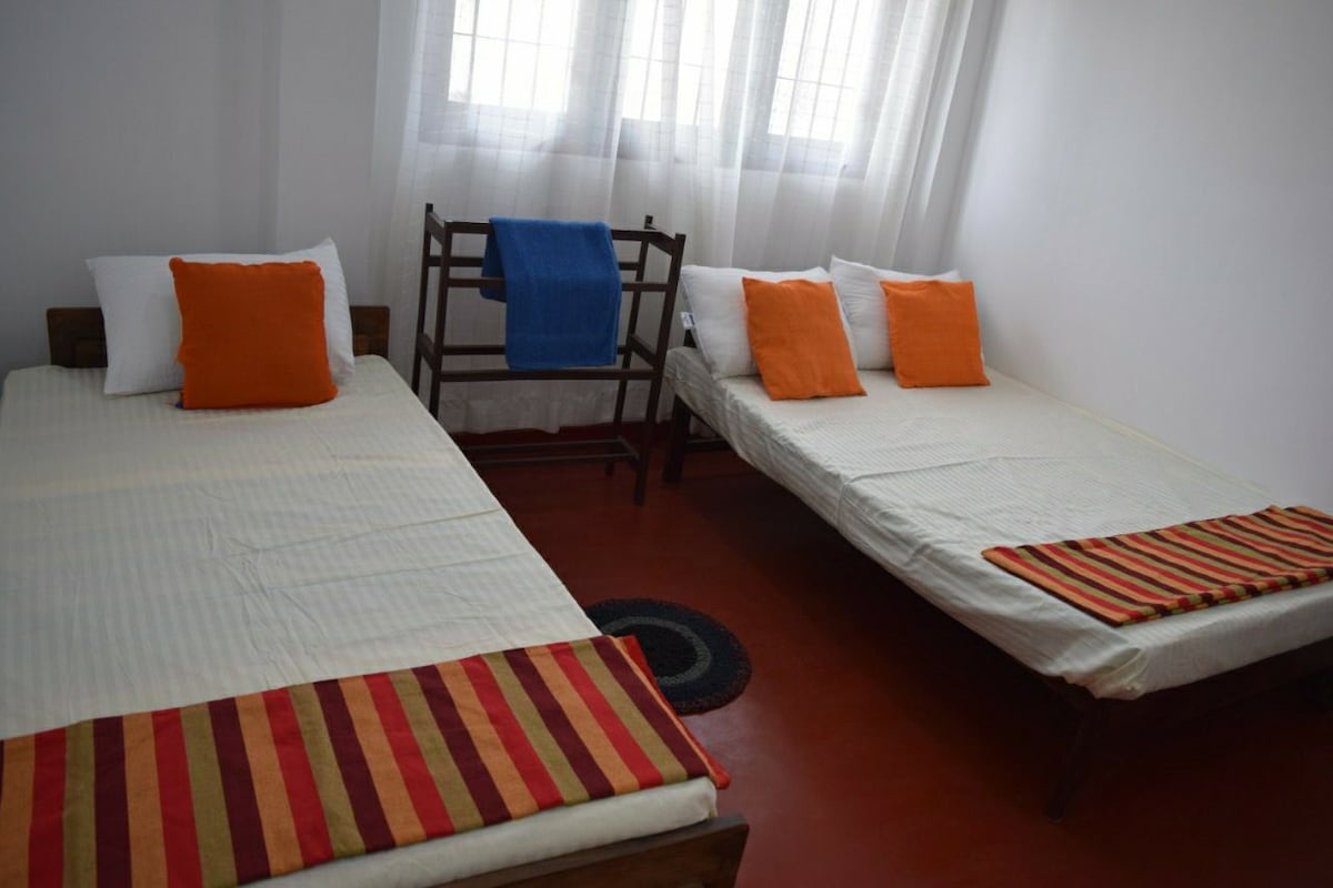 The room features two spacious beds positioned side by side, each adorned with bright orange cushions. A blue blanket is draped over a wooden rack nearby. Large windows allow natural light to fill the room, illuminating the simple, minimalist décor.