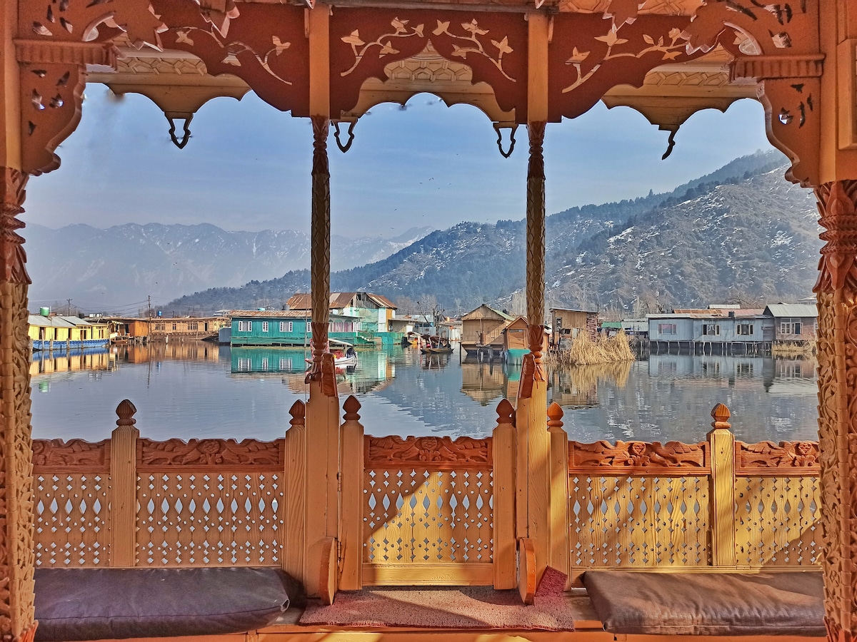 A wooden structure with ornate carvings provides a view of calm waters reflecting nearby houses and mountains. The tranquil scenery includes gentle slopes and a hazy skyline, framed by the intricately designed latticework of the houseboat's seating area.