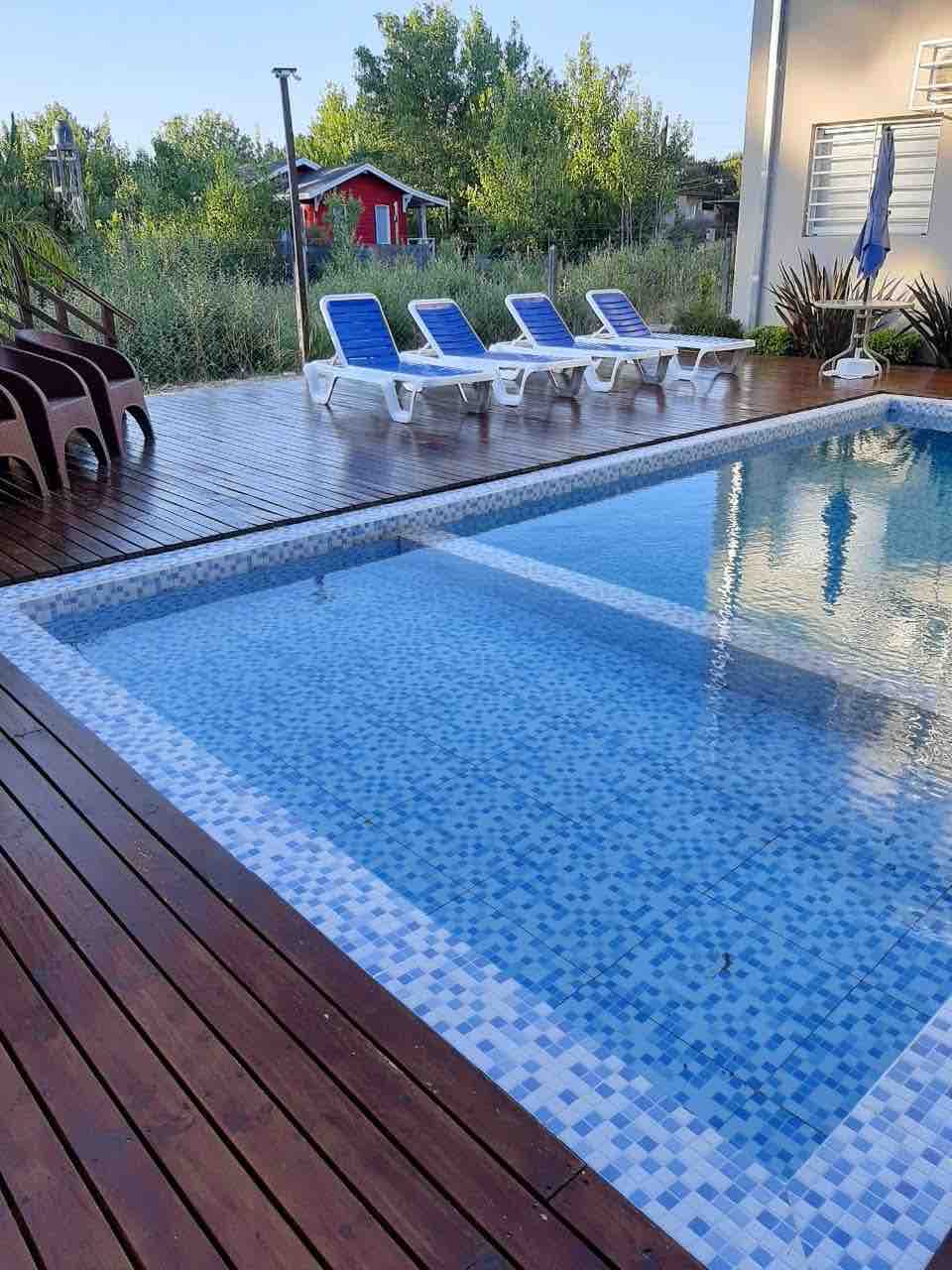 A small shared pool features a tiled border and wooden decking, surrounded by lounge chairs with blue cushions. Lush greenery is visible in the background, enhancing the tranquil atmosphere of the outdoor space.