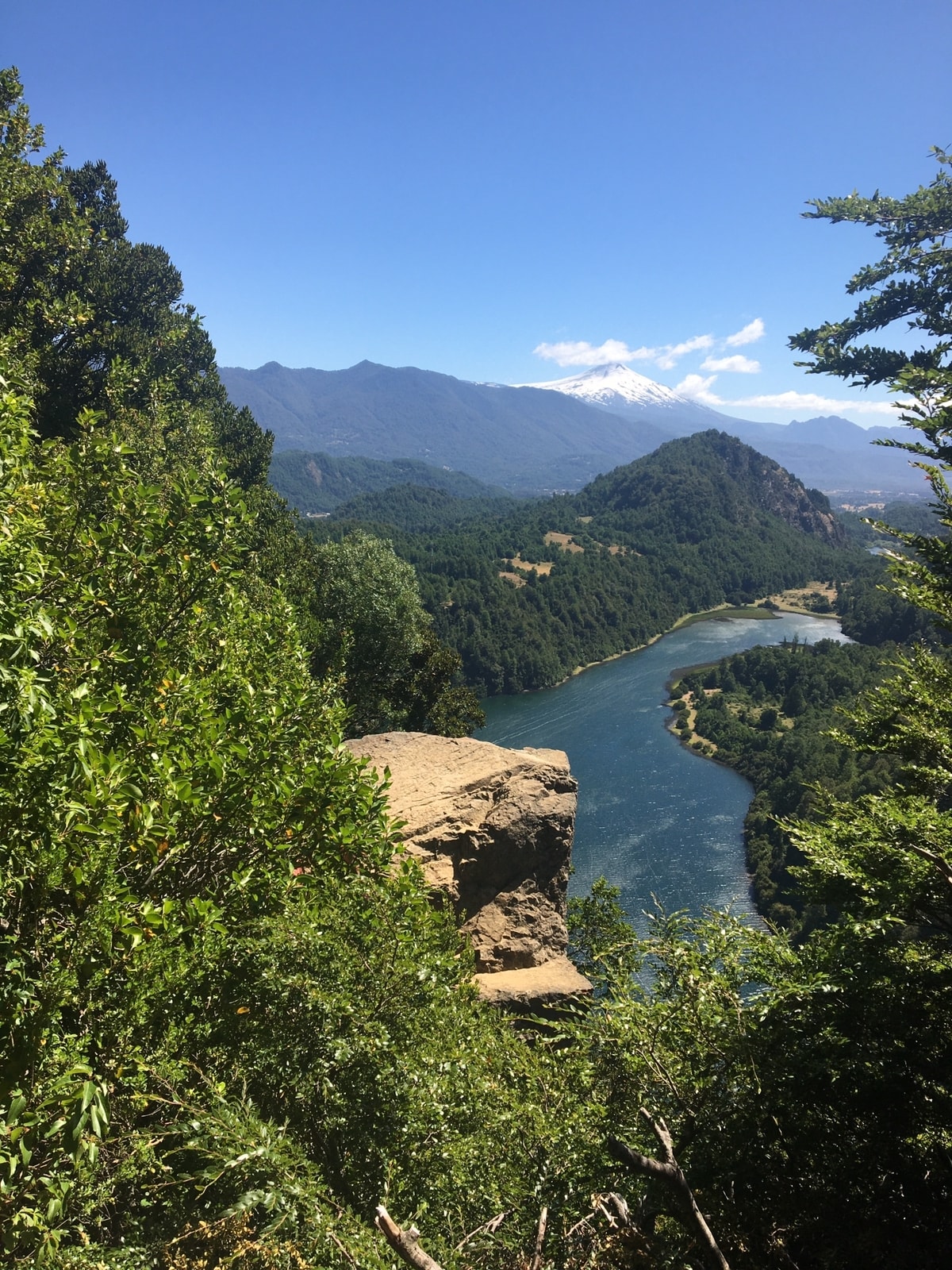 A sweeping view captures a river winding through green valleys, flanked by rocky formations and mountains in the distance. Lush trees and shrubs frame the scene, while a snow-capped peak is visible under clear blue skies.