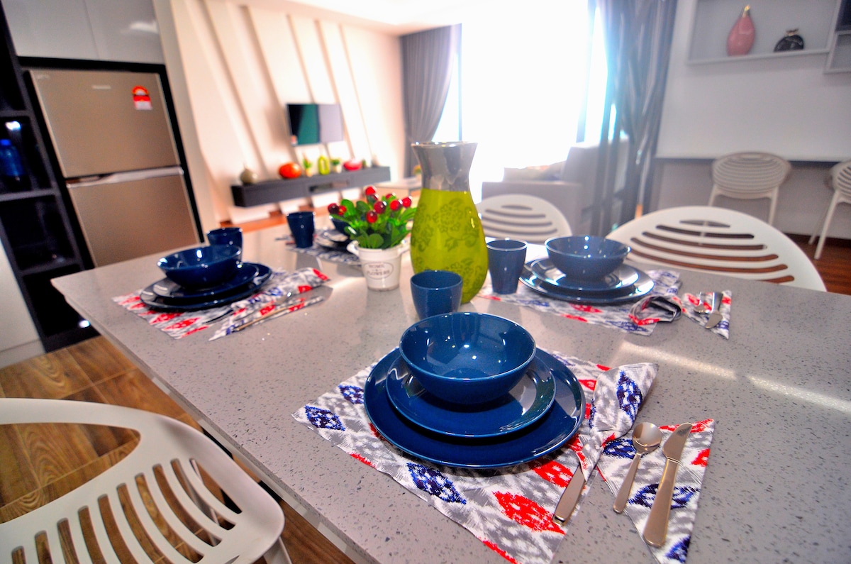 A well-set dining table features an arrangement of deep blue ceramic plates and cups atop patterned placemats. A vibrant green pitcher and a potted plant add color, while a sleek refrigerator and modern living area can be seen in the background.