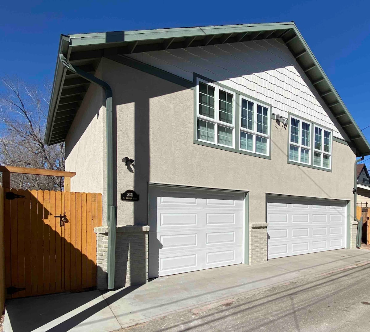 A two-story carriage house facade is displayed, featuring large, symmetrical windows and two garage doors. A wooden fence provides privacy, while blue skies and trees are visible in the background, creating a serene setting.