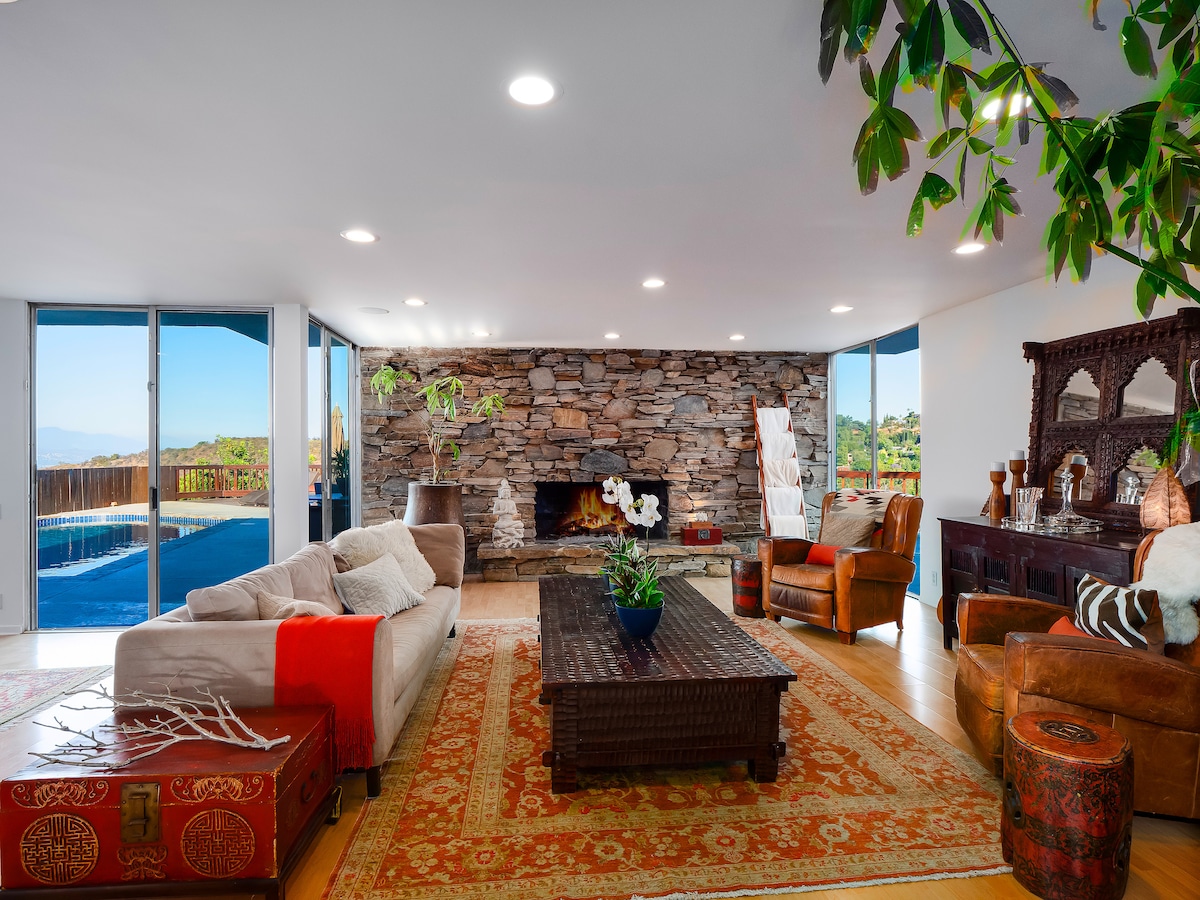 A spacious living area features a stone fireplace as a focal point, surrounded by large windows that offer views of the pool and mountains. Comfortable seating includes a light-colored sofa and warm brown leather chairs. A decorative area rug adds a touch of color, complemented by potted plants.