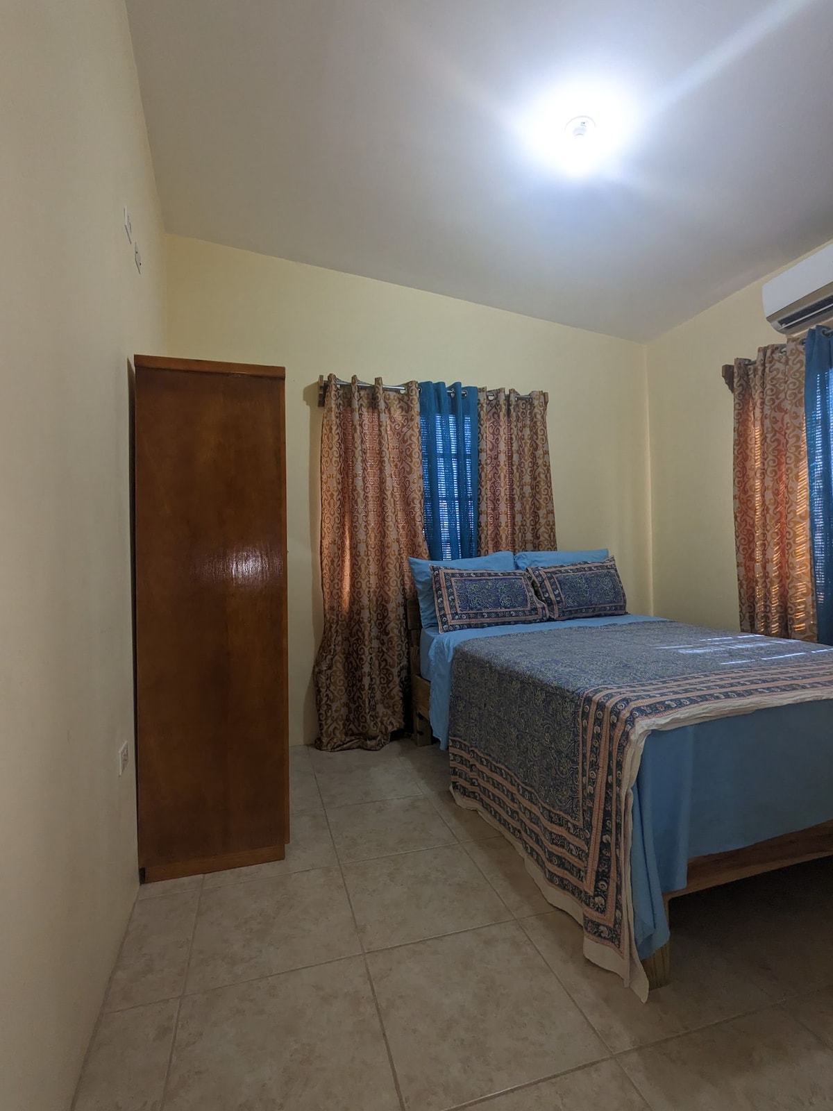 A cozy bedroom is seen with a spacious bed dressed in light blue bedding. Two windows are adorned with patterned curtains that complement the room's warm color palette. A wooden wardrobe is positioned against one wall, enhancing the room's functionality.