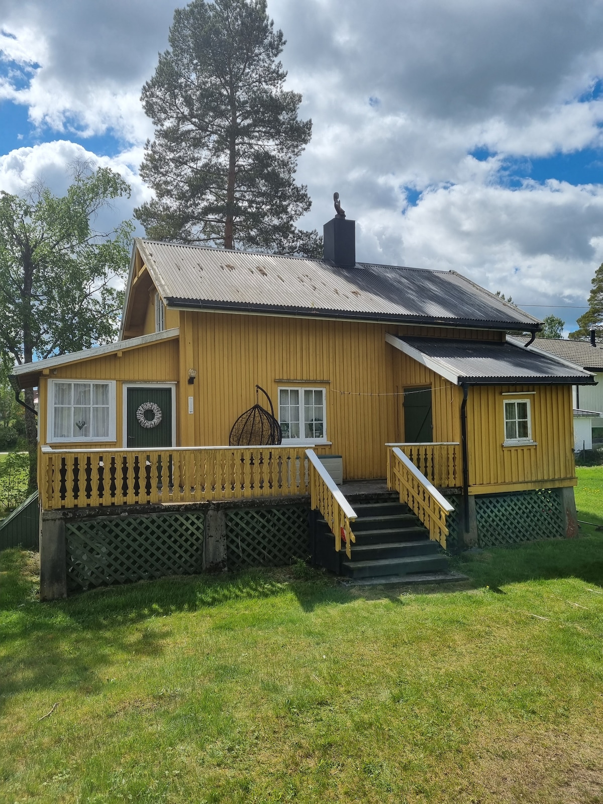A charming yellow wooden cabin stands amidst green surroundings, featuring a sloped roof and a front porch. Stairs lead up to the entrance, while large trees provide shade. The vibrant color and rustic design highlight the cabin's historic charm.